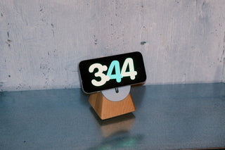 Wooden MagSafe stand holding an iPhone in landscape StandBy mode against a concrete wall.