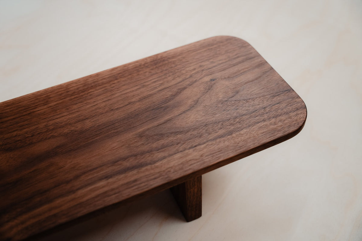 Close-up of the Tilde Monolith monitor riser in solid walnut, showing its rounded corner, continuous grain, and solid walnut leg