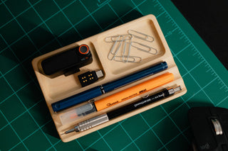 Overhead view of a handmade maple Monolith Desk Tray on a green cutting mat, organizing pens, a mechanical pencil, paper clips, a wireless microphone and a USB-C dongle