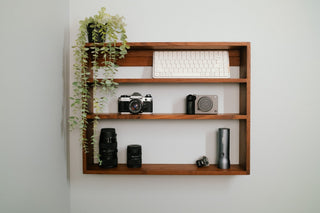 Tilde Made Monolith Wall Shelf in walnut, mounted on a white wall and styled with a film camera, camera lenses, a compact camera, a trailing plant, and an Apple keyboard across three solid hardwood tiers