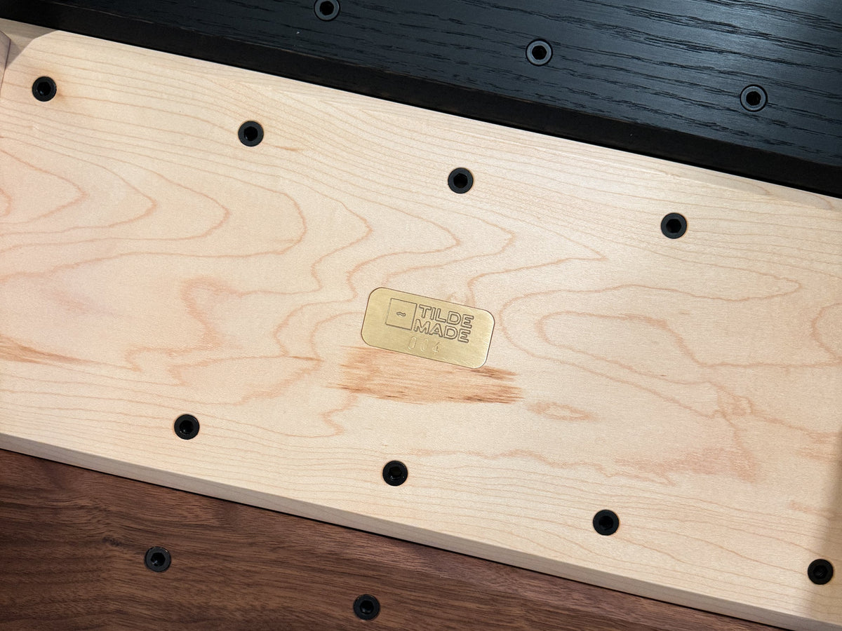 Underside of a solid maple Tilde Monolith monitor riser showing its 1/4"-20 threaded inserts and brass Tilde Made maker's plate, between black oak and walnut risers