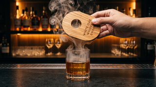 A hand holding the oval White Oak Whiskey Smoker as a lid above a crystal rocks glass of bourbon, oak smoke pouring down into the glass at a moody backlit whiskey bar
