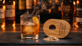 A smoked Old Fashioned with an orange twist and large ice cube beside the oval White Oak Whiskey Smoker and a pile of oak wood chips, smoke curling off the glass in a warm whiskey bar