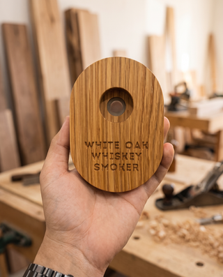 Hand holding a solid white oak whiskey smoker in a woodworking workshop, with stacked lumber and a hand plane in the background