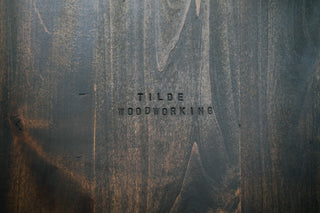 Close-up of dark walnut grain with a 'TILDE WOODWORKING' brand stamp pressed into the wood surface.