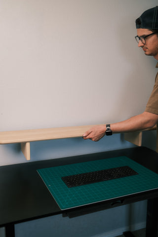 Nicos of Tilde Made placing a maple Monolith Desk Shelf onto a standing desk, shown for scale with a mechanical keyboard and cutting mat.